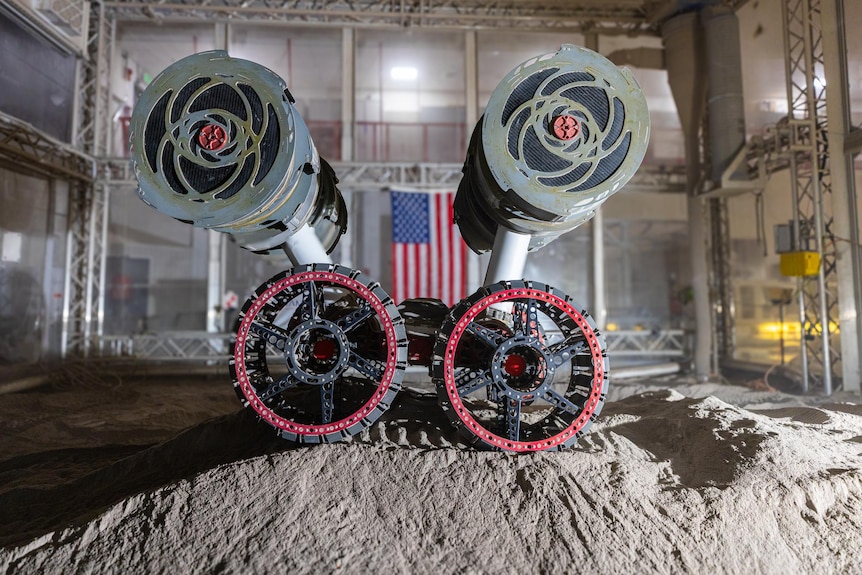 A robot with caterpillar wheels and arms on a mound of dust in front of an American flag.