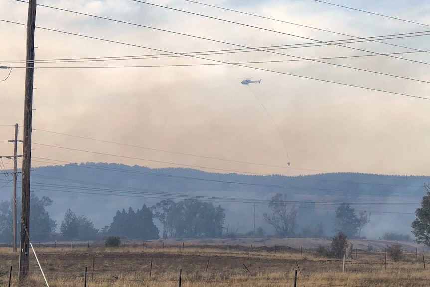 A helicopter flies through smoke, a parachute filled with water trailing below it.