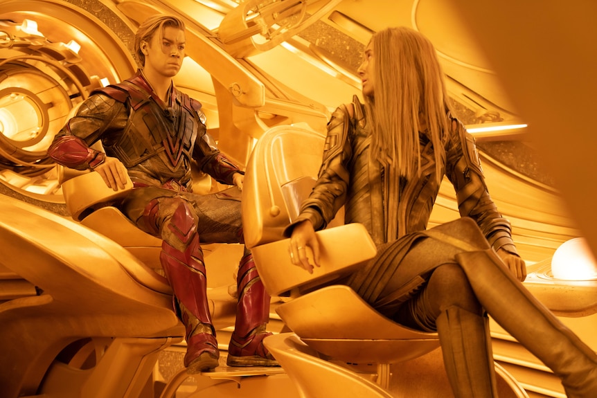 A man and a woman with gold faces and high-tech space suits sit inside a gold-toned space ship.
