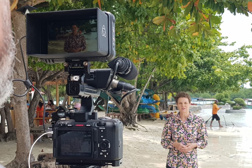 A man operating a camera films a woman standing on a beach talking to camera with people in background walking into water.