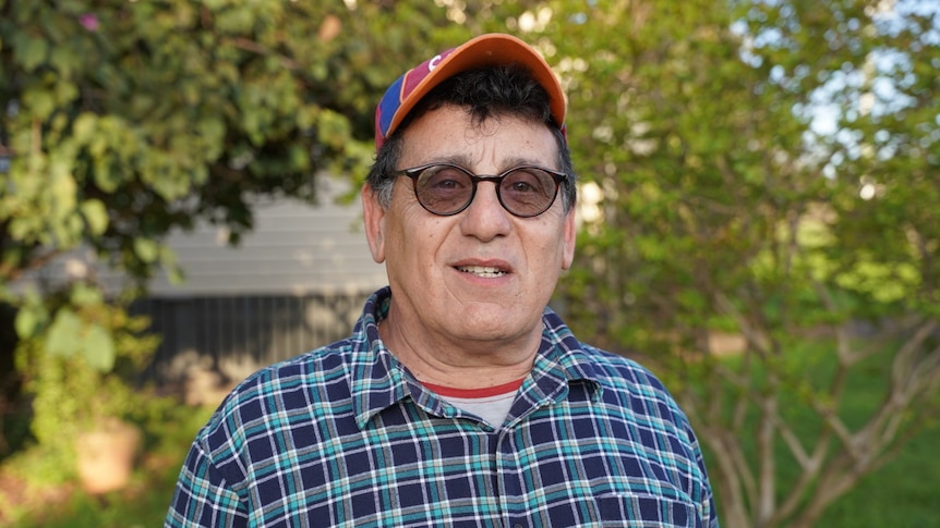 A man wearing a cap and glasses stands outside in front of some greenery.