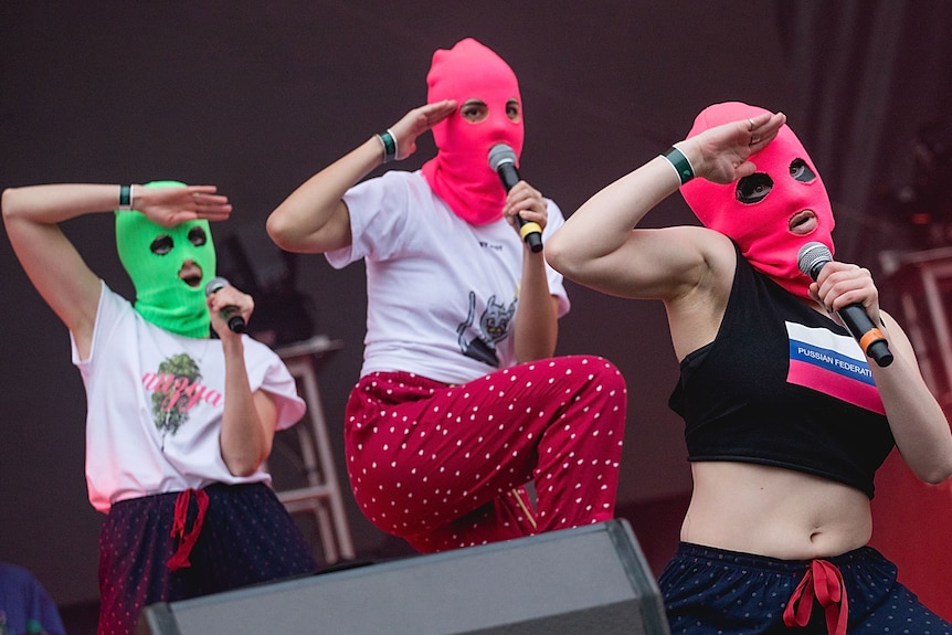 Three people with mics on stage in brightly colored balaclavas
