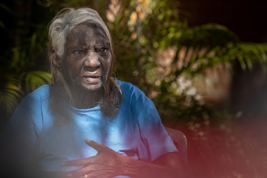 a woman with grey hair sits in front of green palm tree and is looking to the distance. 