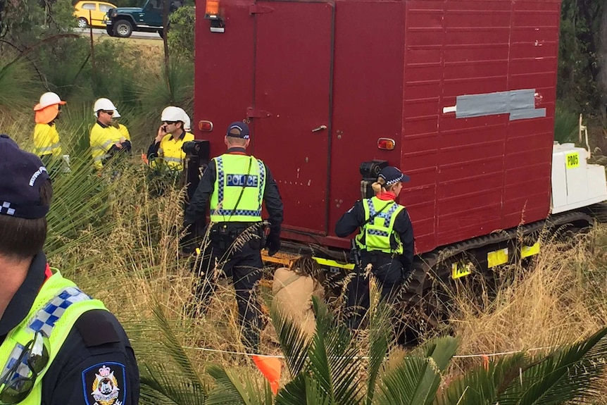 A man kneels at the back of a heavy machine surrounded by police officers and workers.