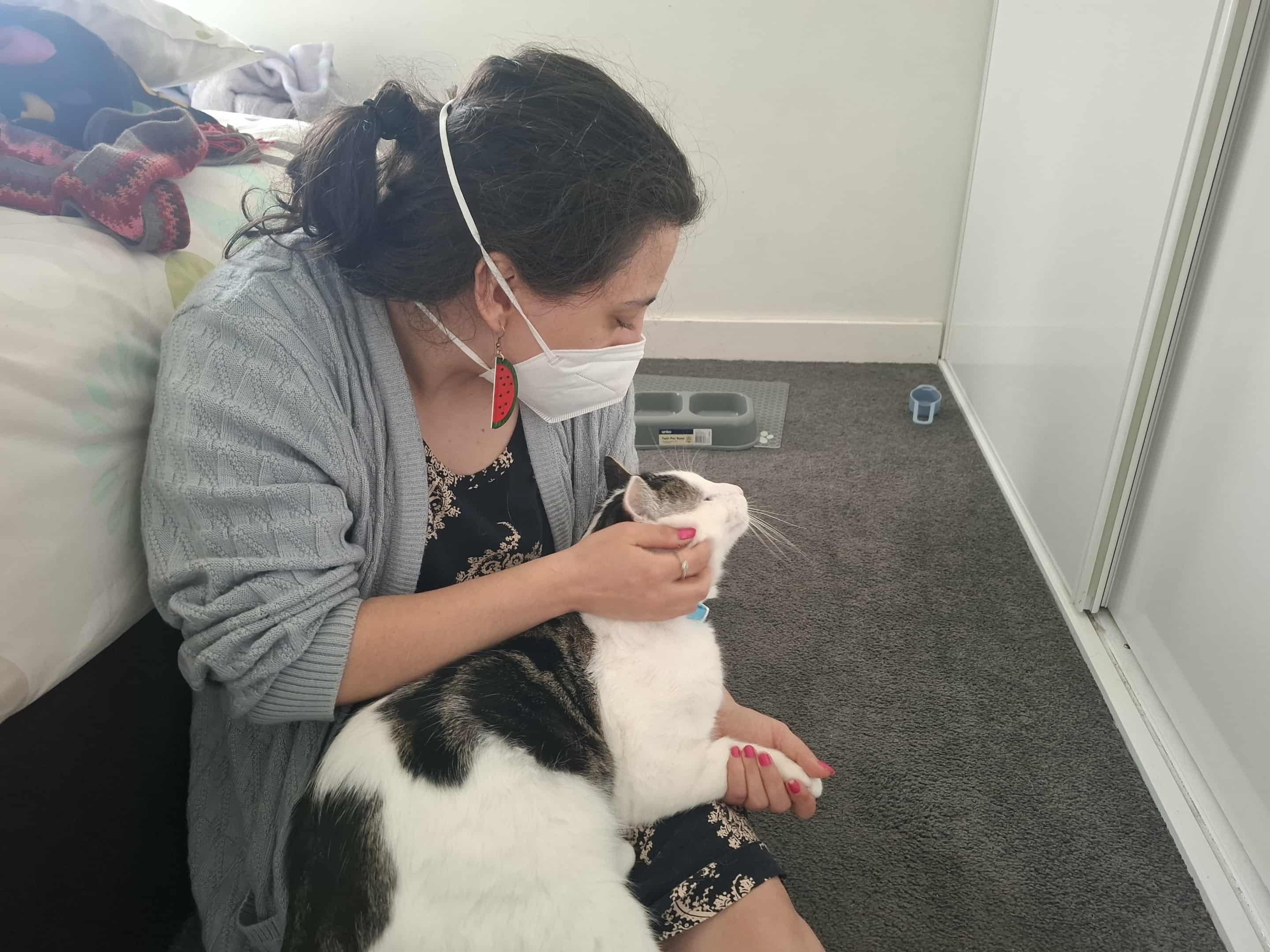 Laura está sentada ao lado da cama usando uma máscara e acariciando seu gato preto e branco, Giles.