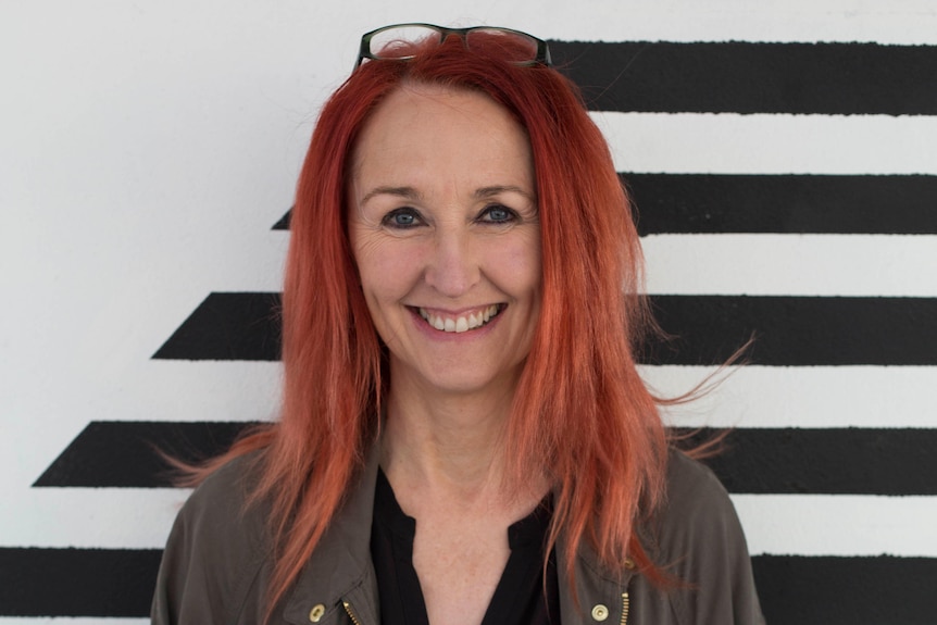 Woman with vibrant red hair wears black shirt and grey jacket and smiles standing against black and white striped wall.