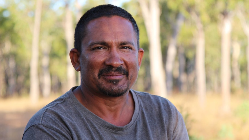 A man in a grey shirt smiles to camera, with an out-of-focus Australian bush background.