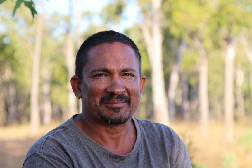 A man in a grey shirt smiles to camera, with an out-of-focus Australian bush background.