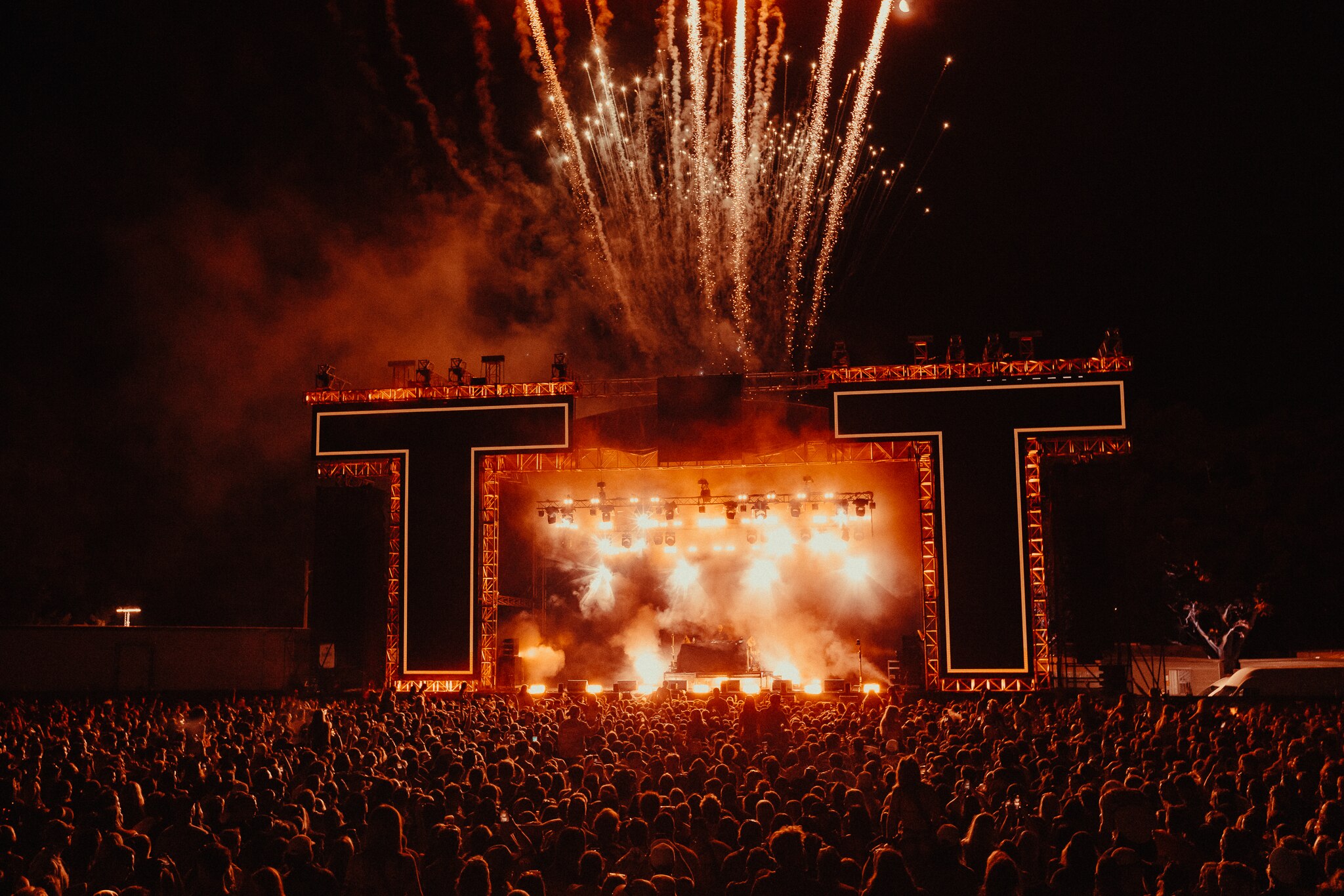 Fireworks and lights let rip at an outdoor stage framed by giant T's and a massive crowd watching at night