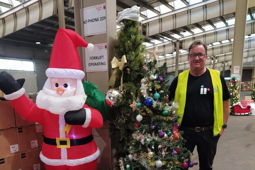 A man stands next to a Christmas tree, plastic blow up Santa and a pile of presents.