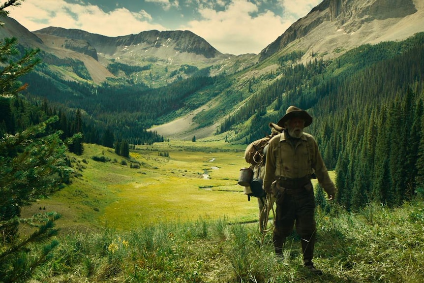 Picture of a green valley, with mountains in background, and scruffy looking older man walking towards camera with horse.