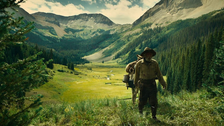 Picture of a green valley, with mountains in background, and scruffy looking older man walking towards camera with horse.