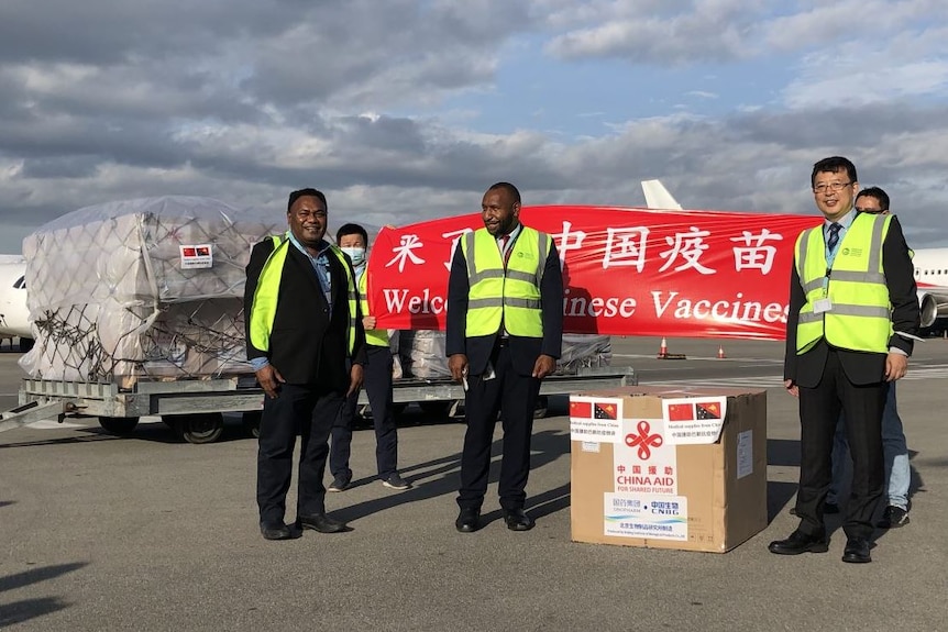 Three men pose near a box of vaccines at the airport with a sign reading 'welcome Chinese vaccines'