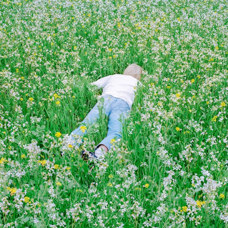 Porter Robinson wearing a white shirt and jeans, laying flat face down in a field of grass and flowers.