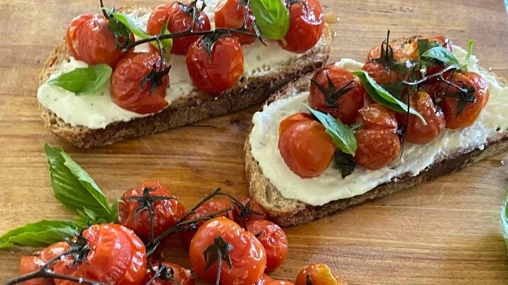 Roasted cherry tomatoes and basil leaves on a wooden board next to pieces of sourdough toast dotted with more of them.