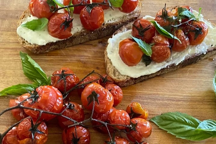 Roasted cherry tomatoes and basil leaves on a wooden board next to pieces of sourdough toast dotted with more of them.