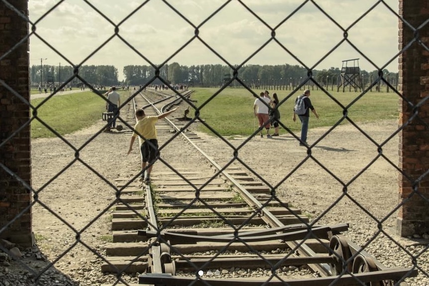 A child, seen from behind through a chain-link fence, balances on the rails of a train track that leads to Auschwitz.