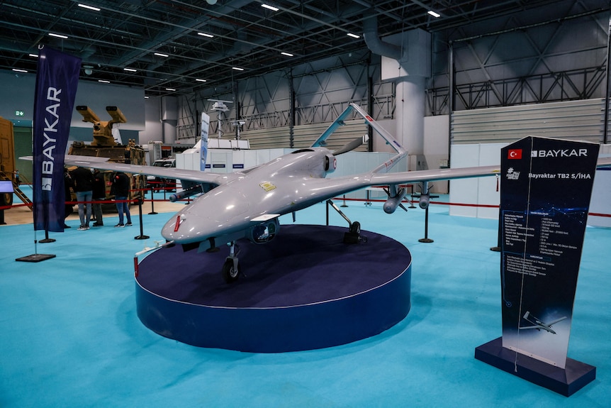 A propeller-driven drone sits atop a small circular stand on display in a large warehouse
