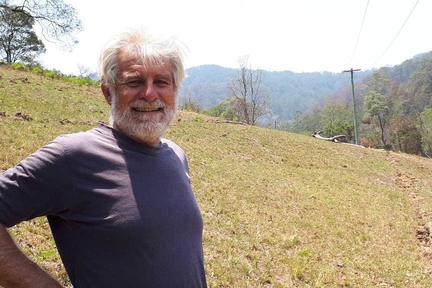 Portrait of Mike Roze smiling, grey hair and beard, standing on a green hill in front of bushland and mountains.