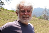 Portrait of Mike Roze smiling, grey hair and beard, standing on a green hill in front of bushland and mountains.