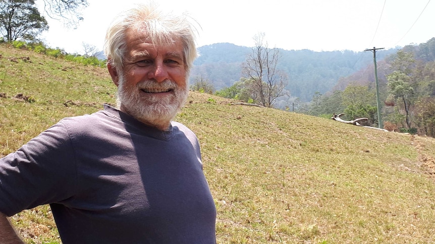 Portrait of Mike Roze smiling, grey hair and beard, standing on a green hill in front of bushland and mountains.