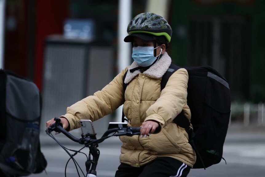 A person riding a bike with a mask in Melbourne. She is wearing a helmet and large backpack for deliveries.