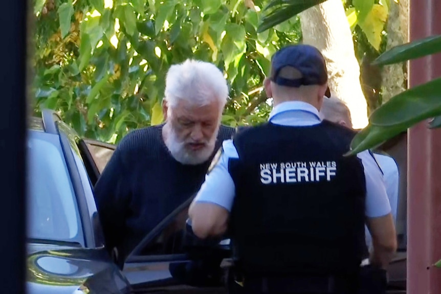 Camera through a gate showing an elderly man with white hair being escorted into a car by police