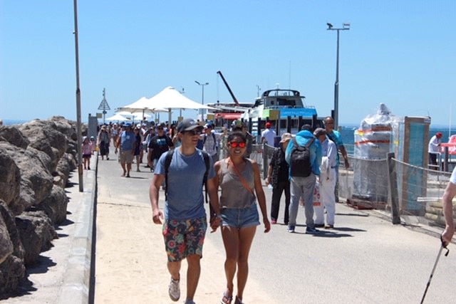 A man and woman hold hands and walk down the Rottnest ferry wharf.