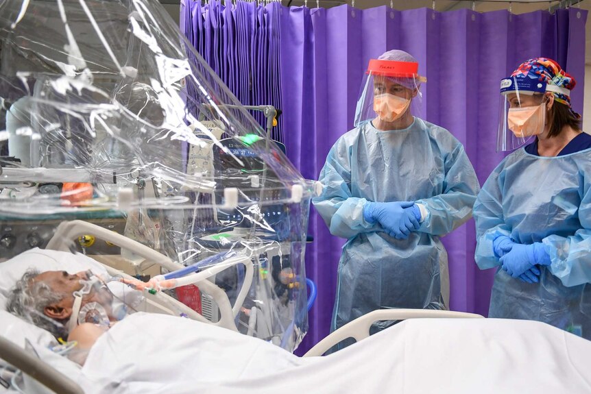 A man lies in an ICU unit with plastic covering his bed. Two people in full PPE gear stand next to his bed watching him.
