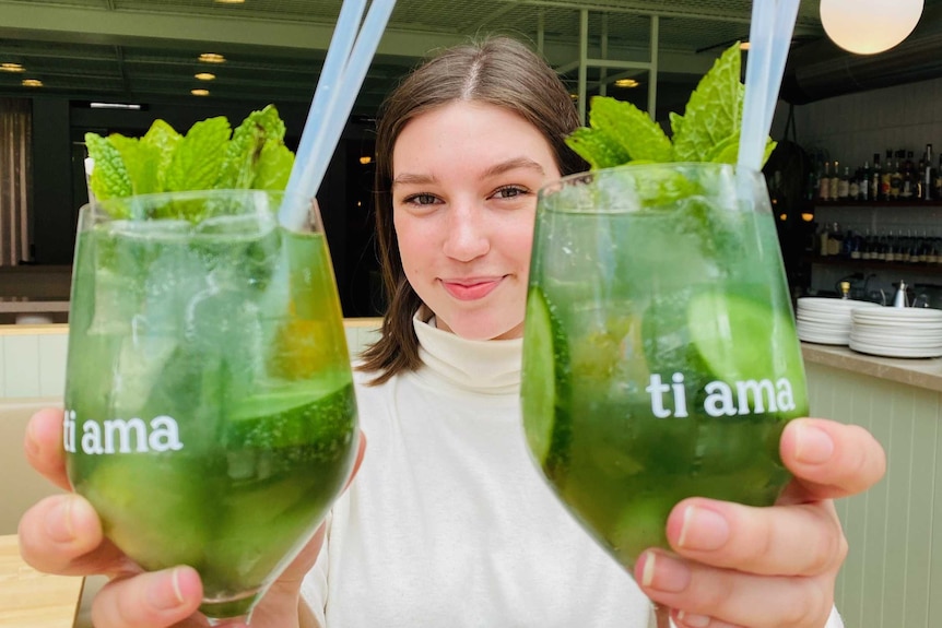 A woman smiles and holds two cocktails with mint in them