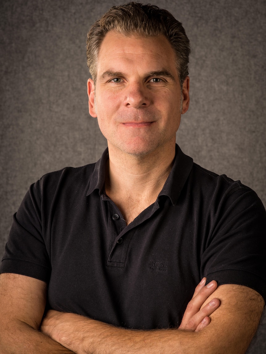 A profile shot of Dean Prenc with arms crossed, black shirt and smile