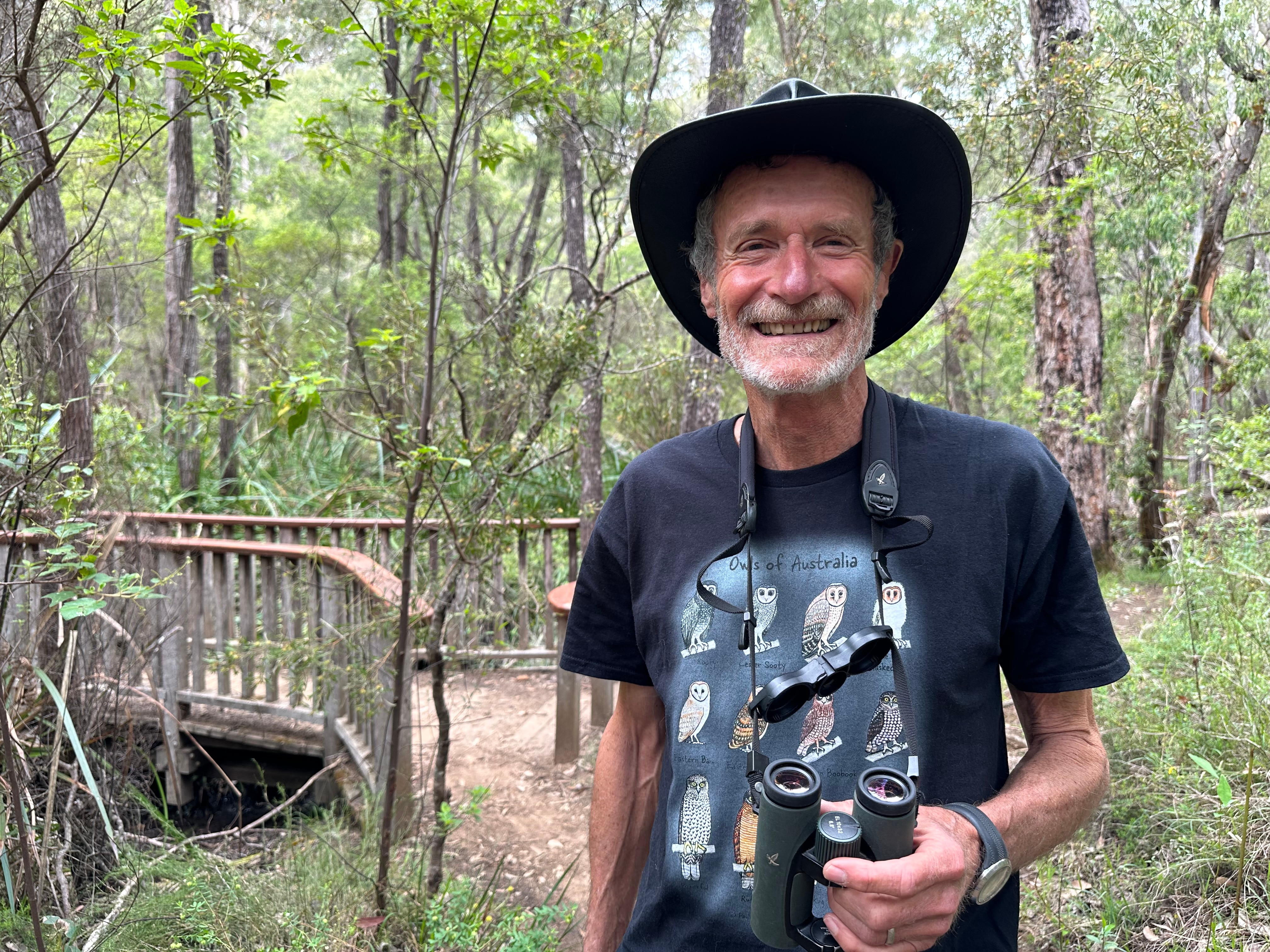 Un hombre de barba corta gris, vestido con una camiseta negra con búhos en el frente y binoculares alrededor del cuello, sonriendo.