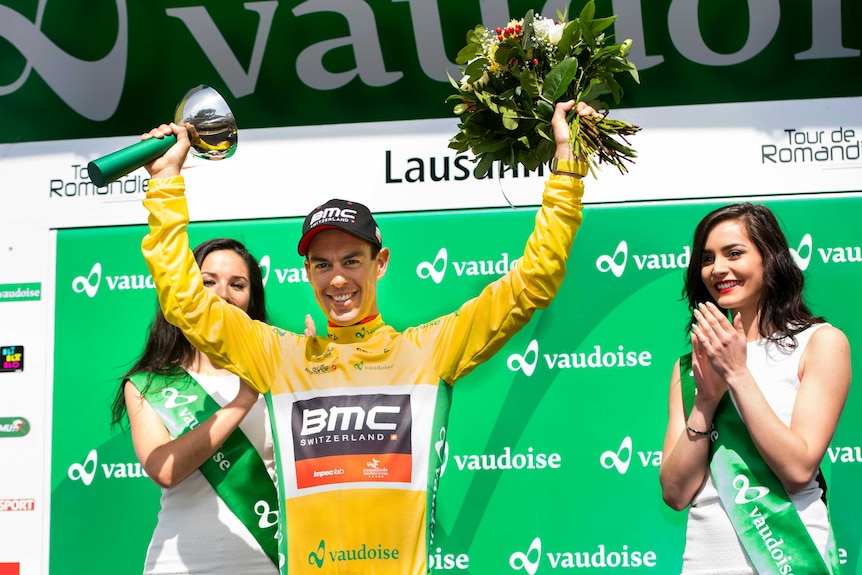 Richie Porte raises his arms with a trophy and flowers after winning a cycling race.