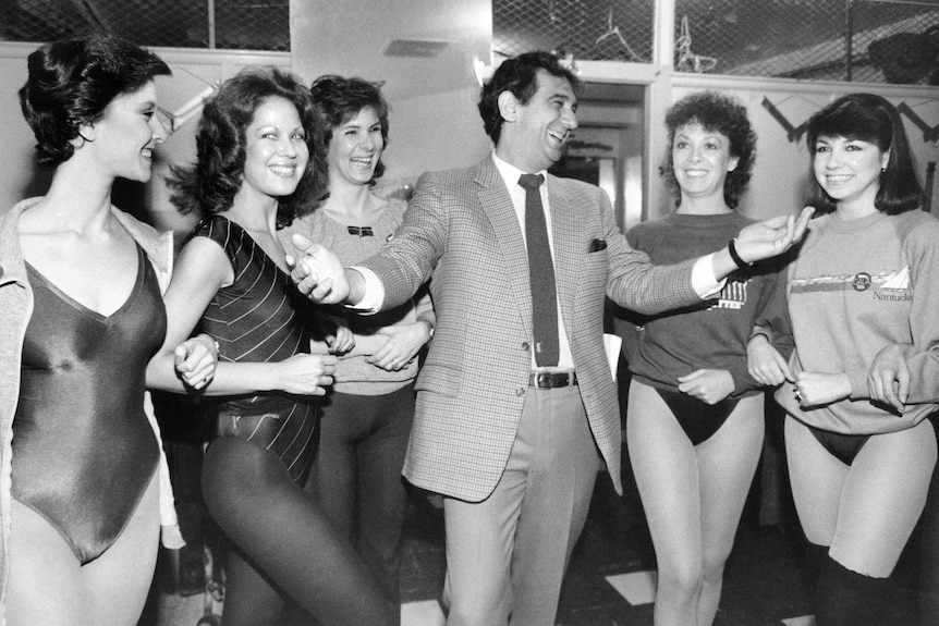 Placido Domingo laughs and holds out his hands surrounded by women.