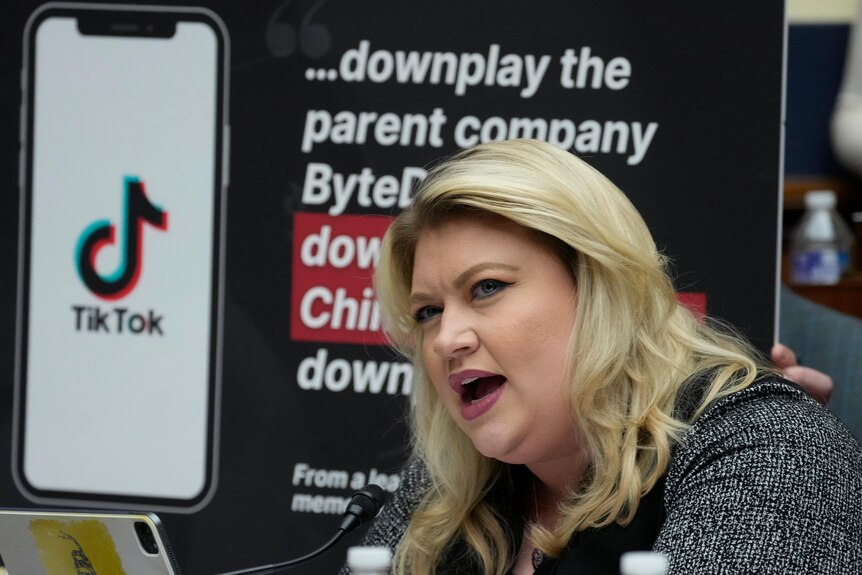 A blonde woman is pictured speaking into a microphone, behind her are placards with the TikTok logo and text.