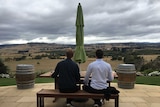 Two people sit on bench with a glass of wine looking out over a rolling paddock