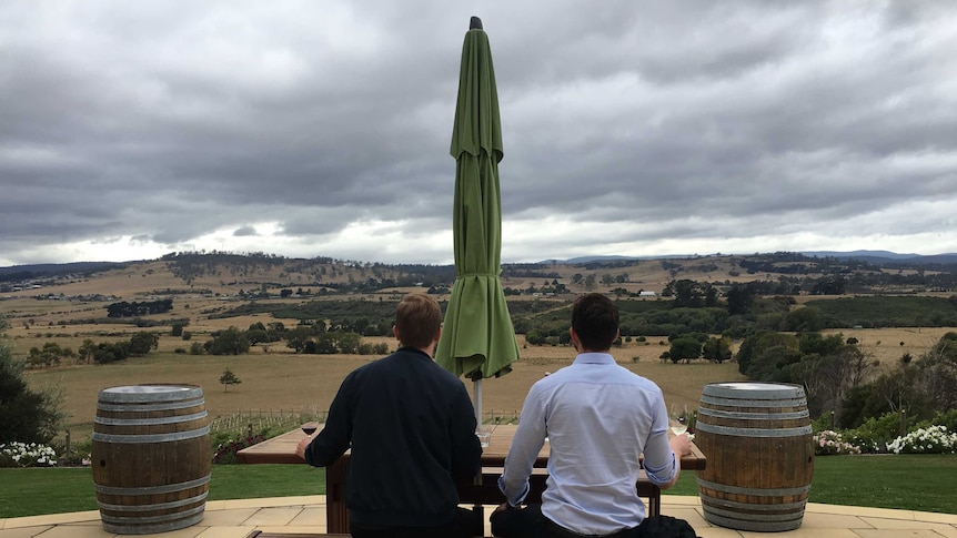 Two people sit on bench with a glass of wine looking out over a rolling paddock