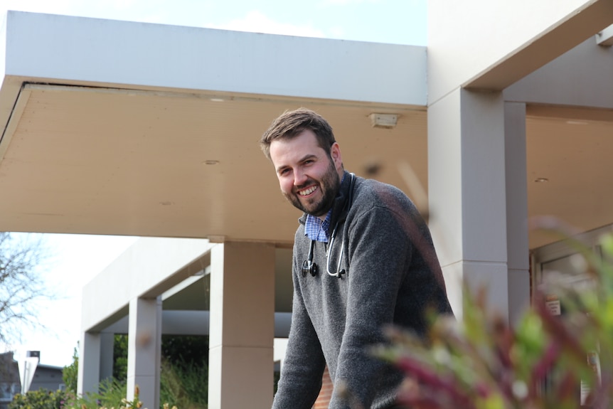 A smiling man with neat dark hair and a beard stands outside a medical clinic. He has a stethoscope around his neck.
