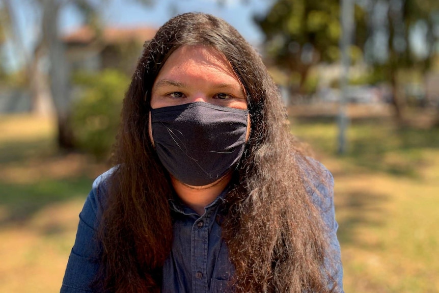 A man sitting in a park, with very long dark brown hair, wearing a black face mask and a navy blue shirt, looks into the camera.