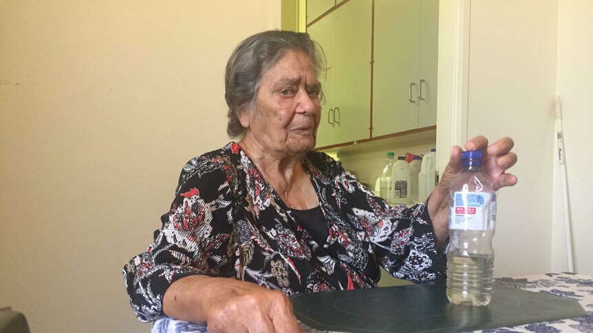 An elderly woman in a kitchen holding a bottle of rainwater.