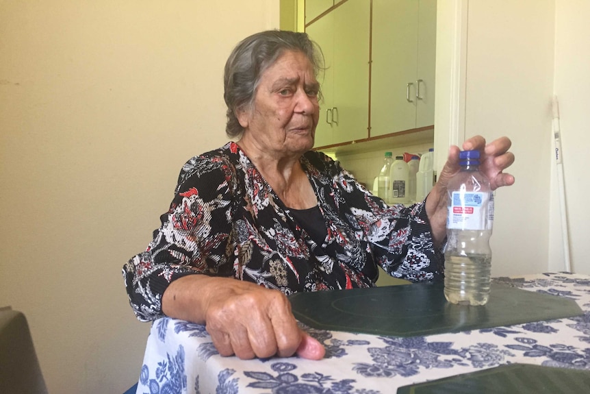 An elderly woman in a kitchen holding a bottle of rainwater.