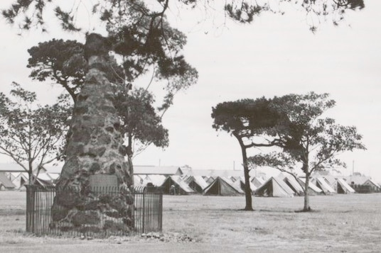 Old black and whiite photo of Burke and Wills memorial, with camp tents in background..