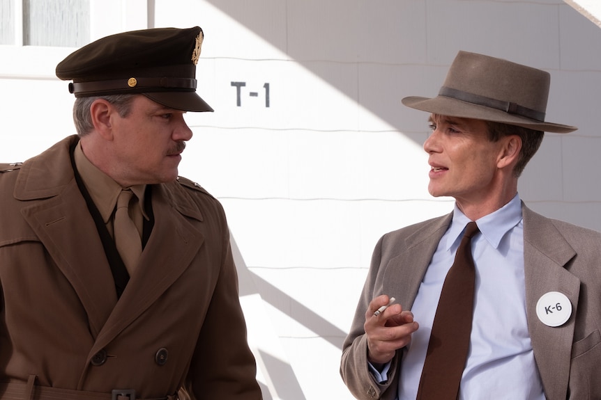 A white man in a tan 40s style American army uniform stands beside another white man in a gray suit with hat and tie.
