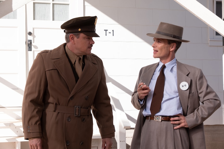 A white man in a tan 40s style American army uniform stands beside another white man in a gray suit with hat and tie.