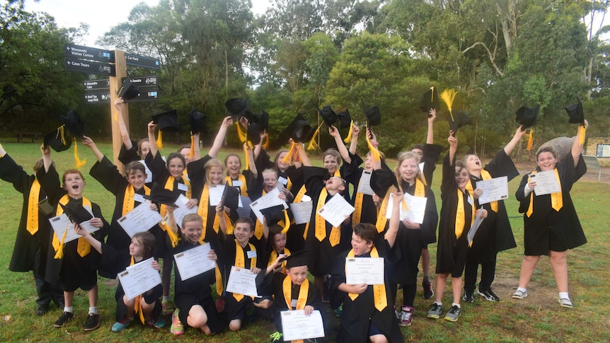 A group of children in black gowns through their mortar-boards in the air in celebration. They are holding certificates.