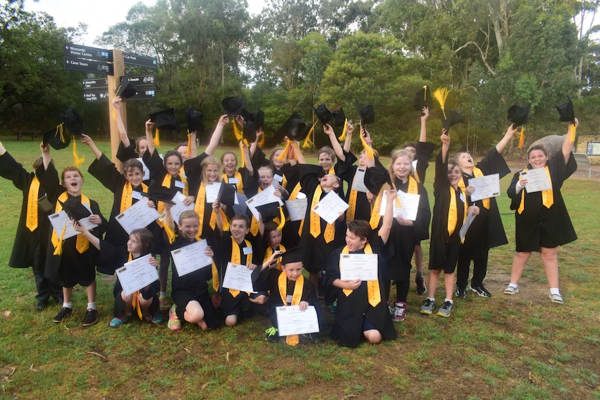 A group of children in black gowns through their mortar-boards in the air in celebration. They are holding certificates.