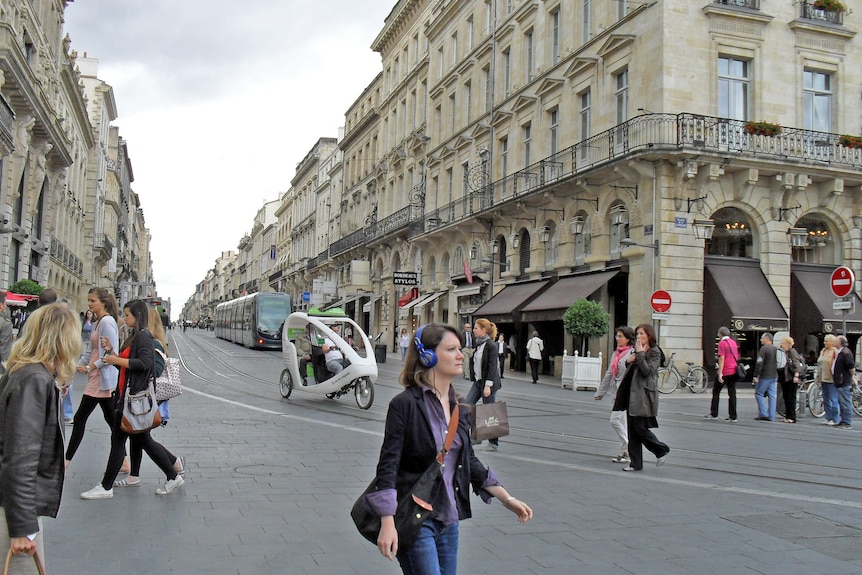 People walking along a busy Paris street, a tram in the background and a woman wearing headphones in the foreground