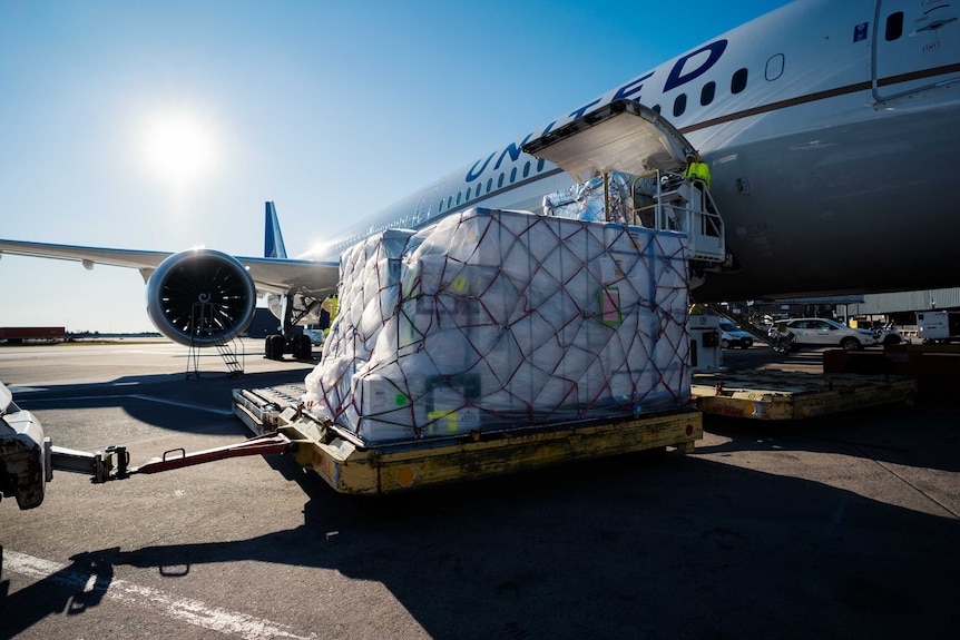 A container being pulled by a car sits on the tarmac with a United airplane behind it.