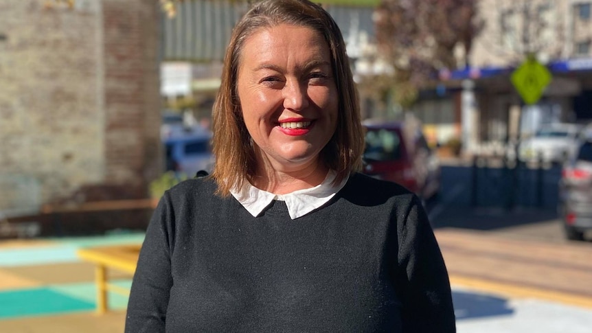 A woman with tawny shoulder-length hair standing outside in the sun with a big smile on her face.