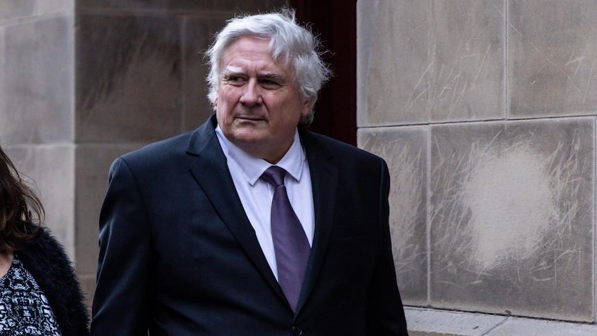 A man with grey hair wearing a black suit and purple tie.